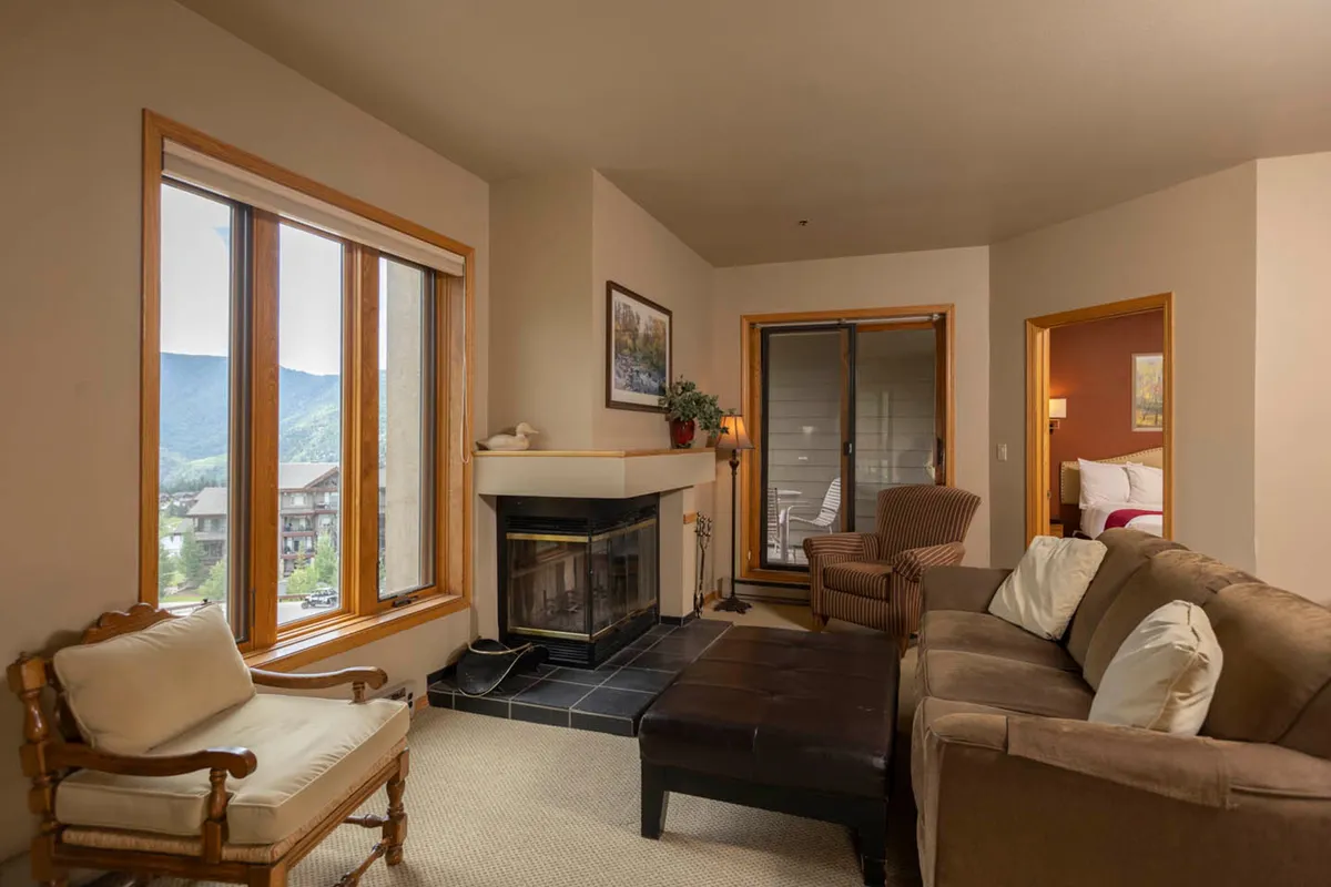 This image showcases a warm, inviting living room featuring a central wood-burning fireplace surrounded by plush, comfortable seating. Expansive windows let in natural light and offer views of the mountains, while the intimate arrangement encourages relaxation and conversation.