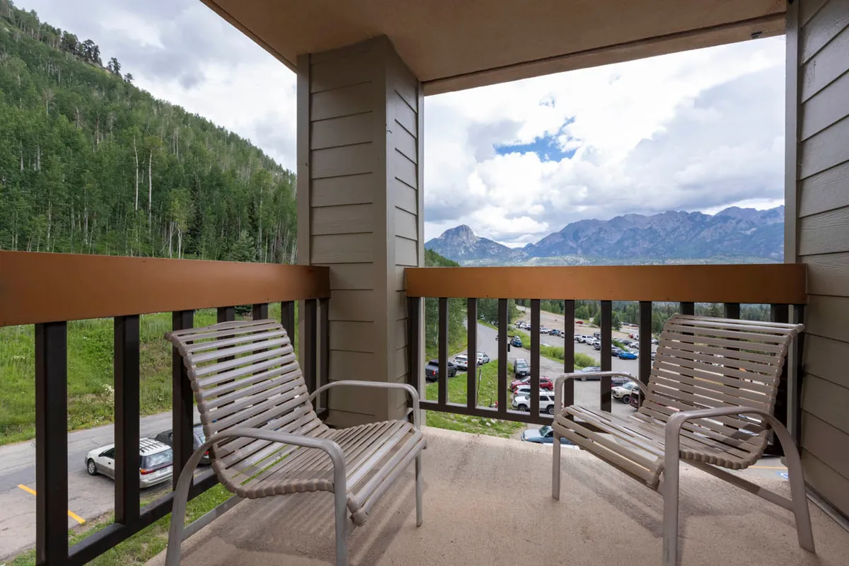 This tranquil exterior shot shows a private balcony furnished with comfortable chairs, set against a dramatic mountain backdrop and lush tree line. The inviting setup is perfect for couples to unwind together, enjoy the fresh alpine air, or simply take in the breathtaking sunrise or starlit sky.