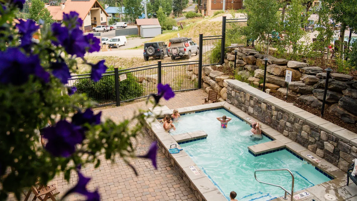 A lively outdoor pool area is shown with people swimming, lounging, and socializing. The pool is surrounded by tasteful landscaping and seating, with the sense of community and leisure palpable in the bright, sunny ambiance.