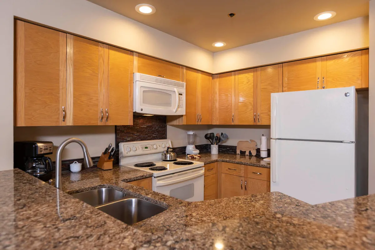 A functional kitchen scene featuring a granite countertop, warm wooden cabinets, white appliances, and a sink. The layout emphasizes practicality and comfort, suitable for preparing everything from pancake breakfasts to romantic dinners.