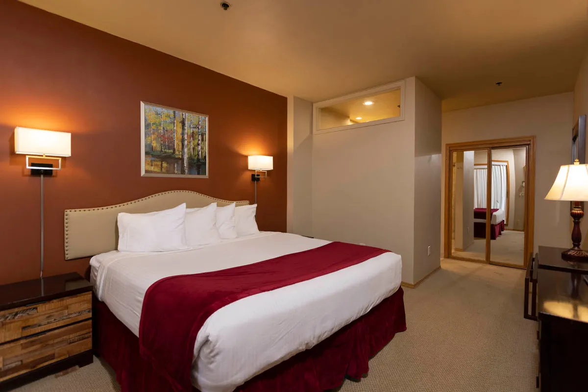 A tranquil bedroom scene featuring a cozy bed with plush bedding, a nightstand with a lamp, a mirror, and tasteful artwork. Soft ambient lighting and neutral tones dominate the space, creating a restful atmosphere ideal for relaxation.