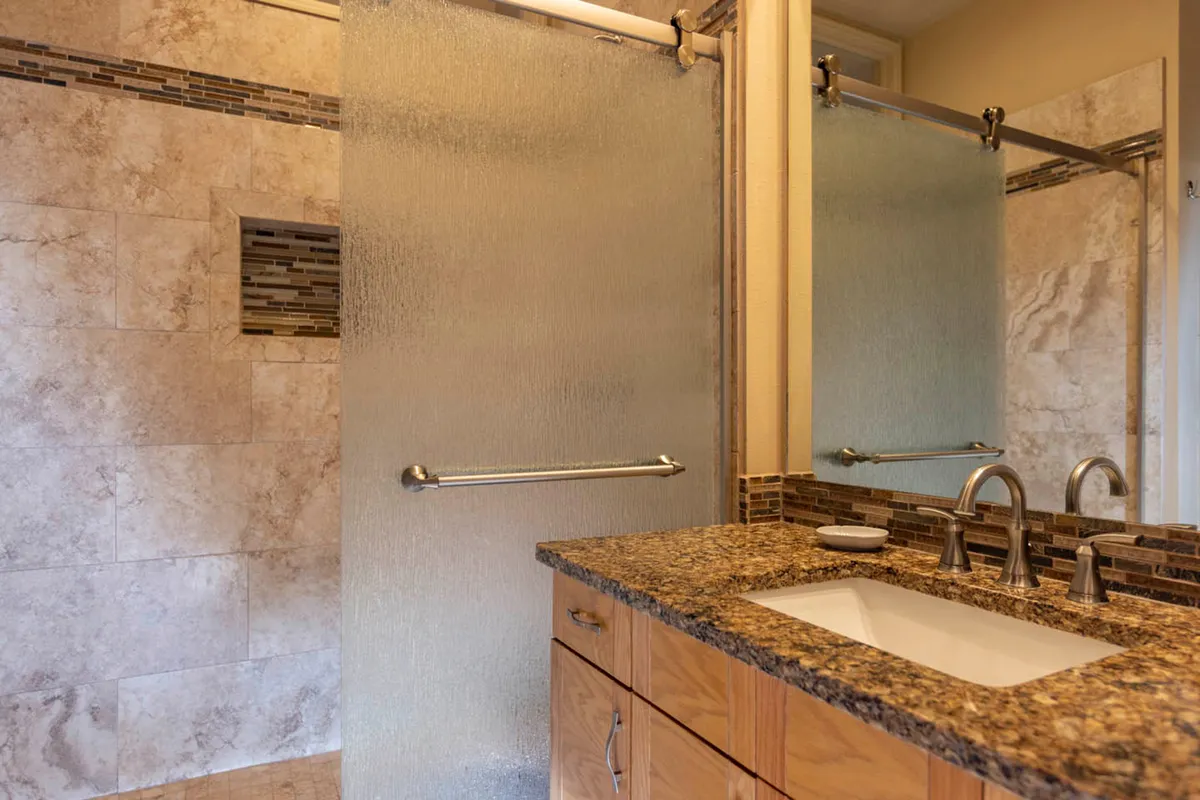 A modern bathroom interior with a granite countertop, glass shower door, sleek faucet, and textured tile wall. The environment is clean, serene, and well-lit—an inviting space for self-care and refreshing routines.