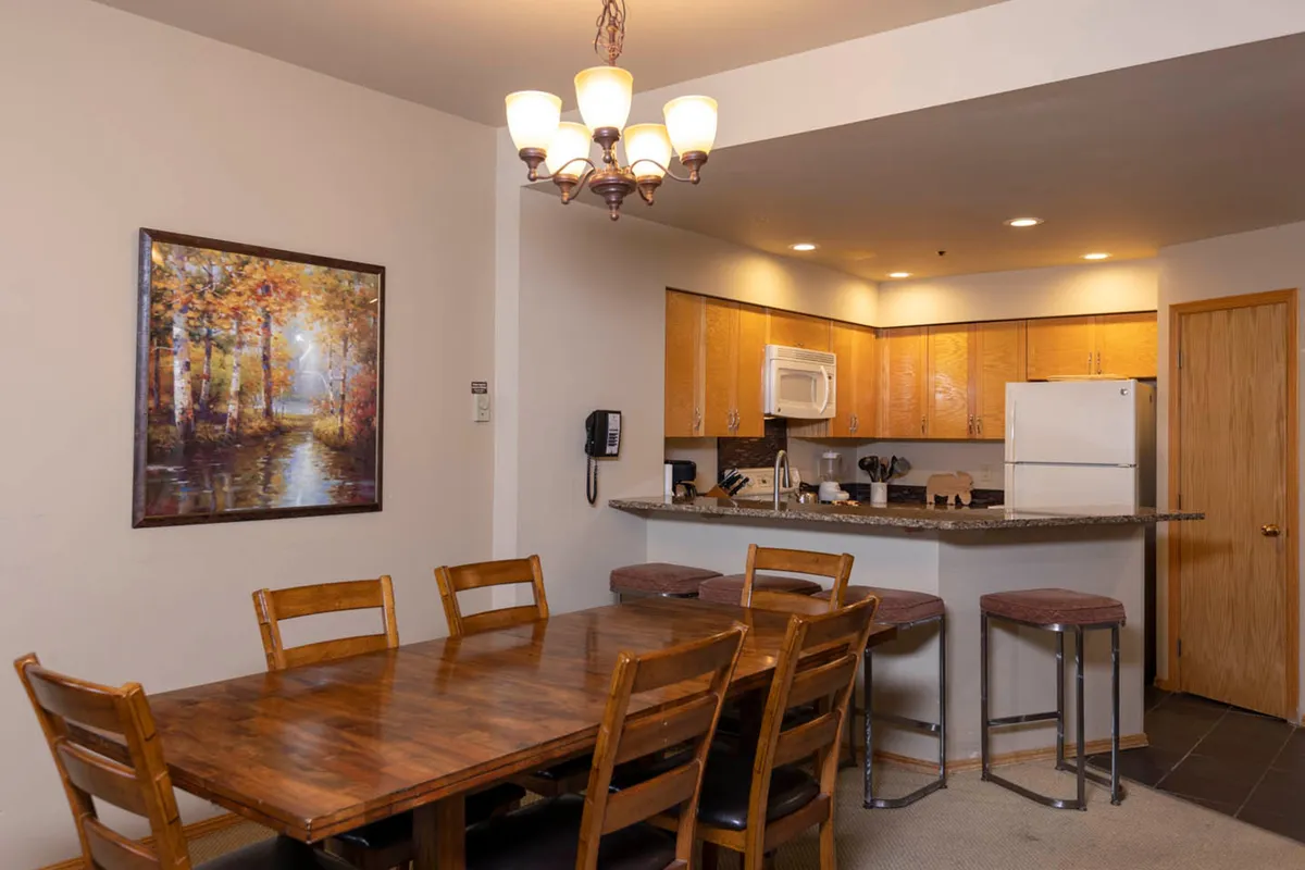 A warmly lit, welcoming dining area with a central dining table set beneath a chandelier, adjacent to a spacious kitchen countertop and framed artwork on the wall. The room is styled for intimate gatherings and family meals, offering a cozy atmosphere that encourages conversation and connection.
