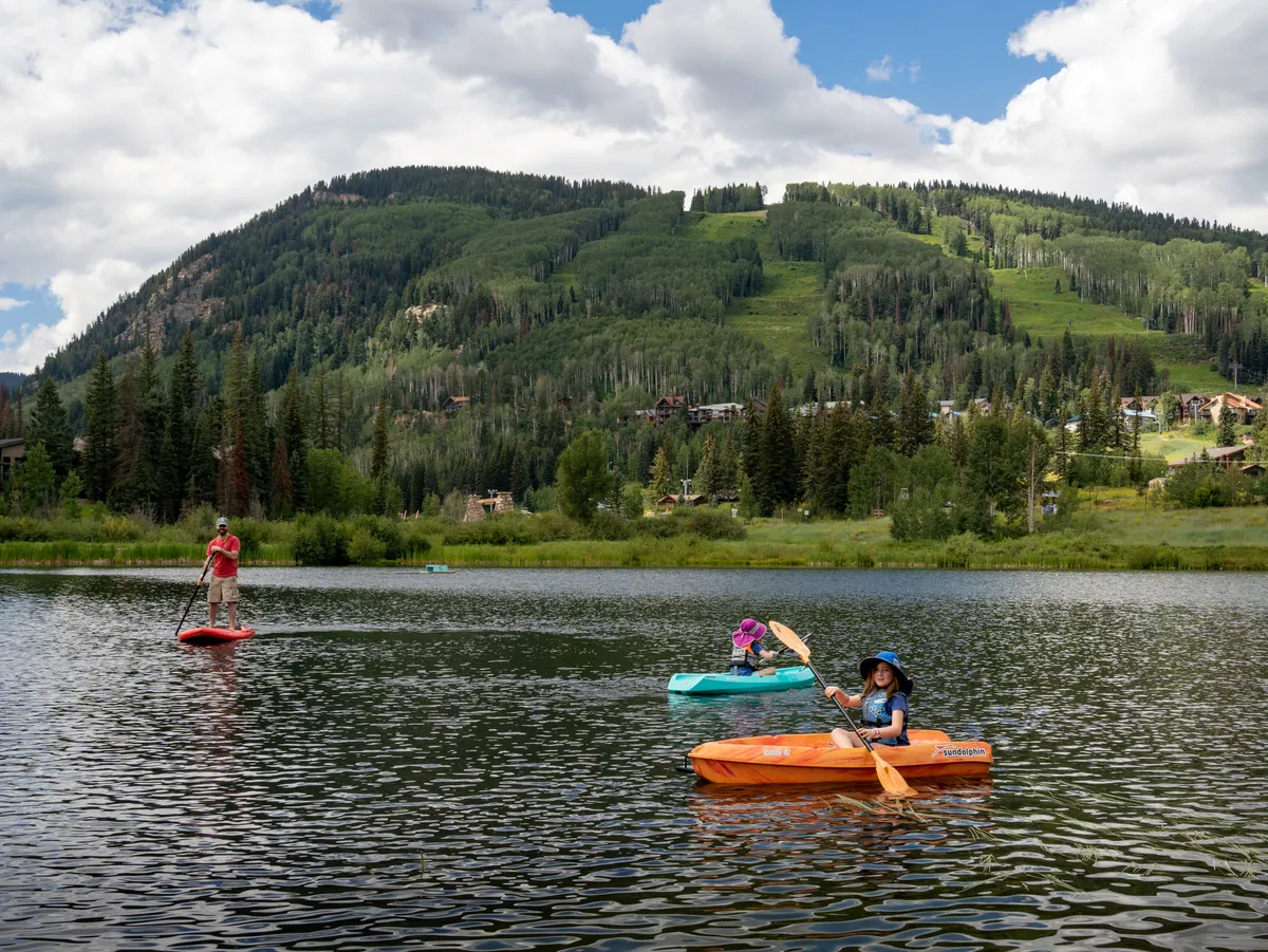 A group of paddleboarders and kayakers enjoy a tranquil lake surrounded by mountains, with clear reflections on the water and an open, active vibe. The mood is joyful and communal, highlighting an adventurous day out in nature.