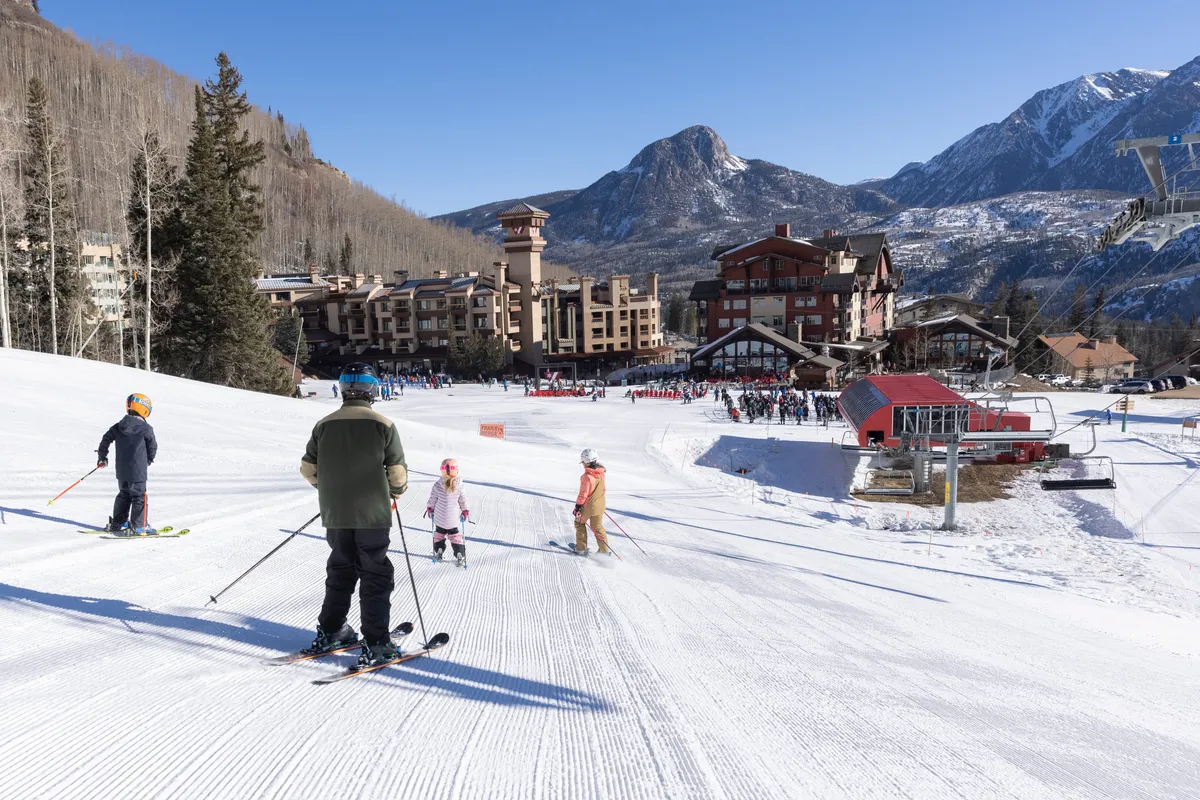 This image shows families and friends enjoying the ski slopes with a scenic mountain backdrop and a ski lodge in view. The atmosphere is lively and adventurous, capturing people learning to ski, snowboarding, and socializing at the base of the slopes.