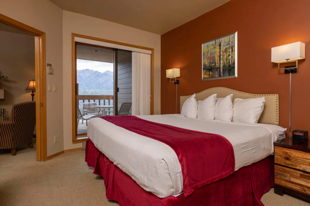 A spacious bedroom with a large bed dressed in decorative pillows, a piece of art on the wall, and a chair by a sliding door that opens to mountain views. The scene exudes warmth and coziness, blending interior comfort with a hint of the surrounding scenery.