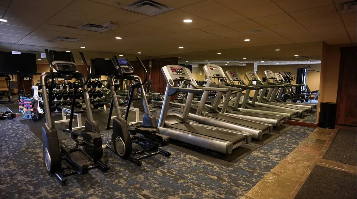 A bright, modern fitness center equipped with treadmills, elliptical machines, and a dumbbell rack. The space is designed to encourage both solo workouts and group fitness activities, with large windows that may offer glimpses of the mountain environment.