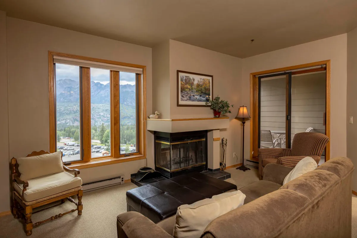 The image showcases a warm and inviting living area featuring large windows framing breathtaking mountain views, a crackling wood-burning fireplace, plush seating, and thoughtfully chosen decorative artwork. The tranquil setting invites guests to unwind, whether watching a sunset, curling up with a book by the fire, or simply soaking in the serene alpine ambiance.