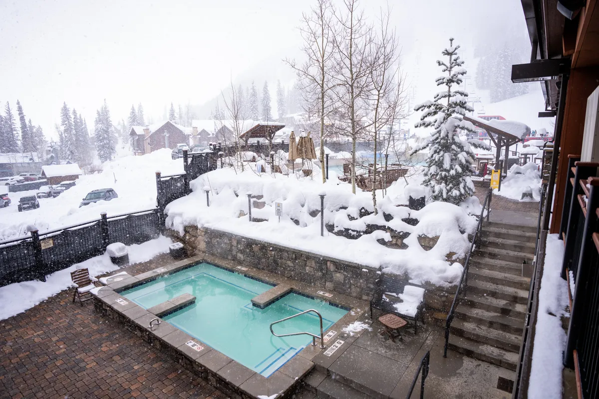 This exterior photo captures a steaming hot tub nestled amidst a snow-covered landscape, surrounded by trees and a staircase leading up to the rooftop area. The inviting scene features people relaxing in the hot tub, soaking up the warmth while gazing out onto the wintry scenery.