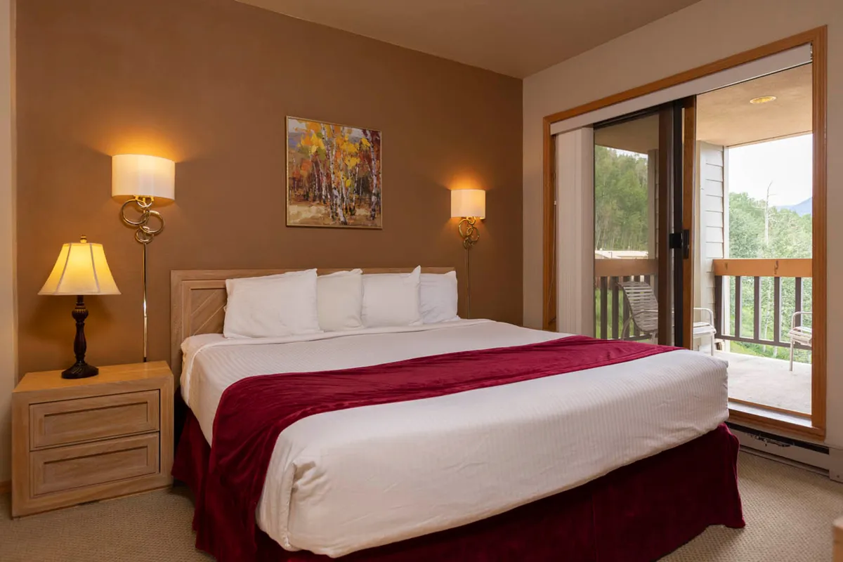 A cozy, well-lit bedroom interior featuring a comfortable bed, stylish artwork, a bedside lamp, and a balcony door that hints at access to the outdoors. The scene suggests relaxation opportunities, enjoying the view, or sipping coffee while starting the day.